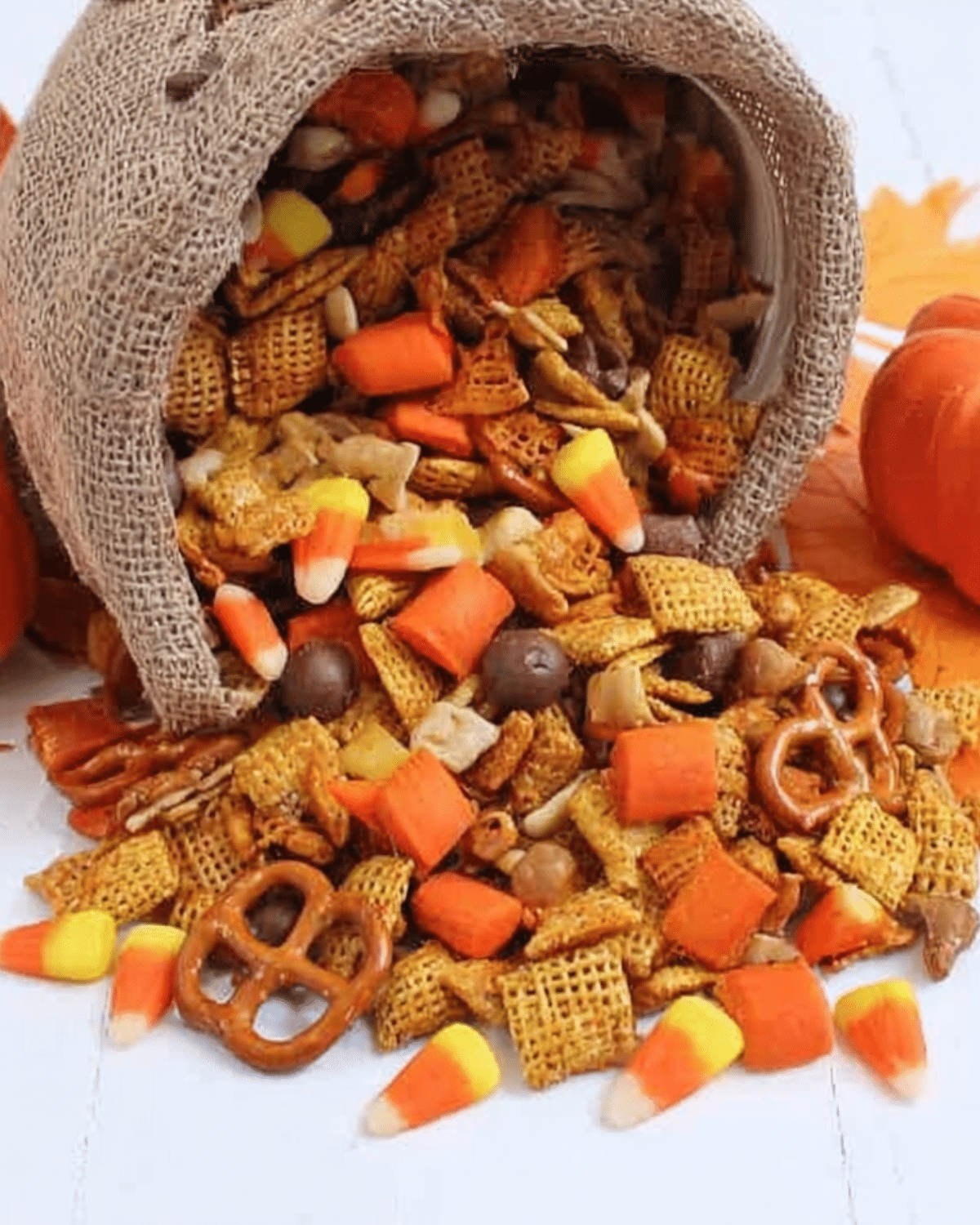 A festive assortment of Pumpkin Spice Snack Mix spilling out of a burlap-wrapped container, featuring candy corn, cereal squares, pretzels, and fall-themed sweets.