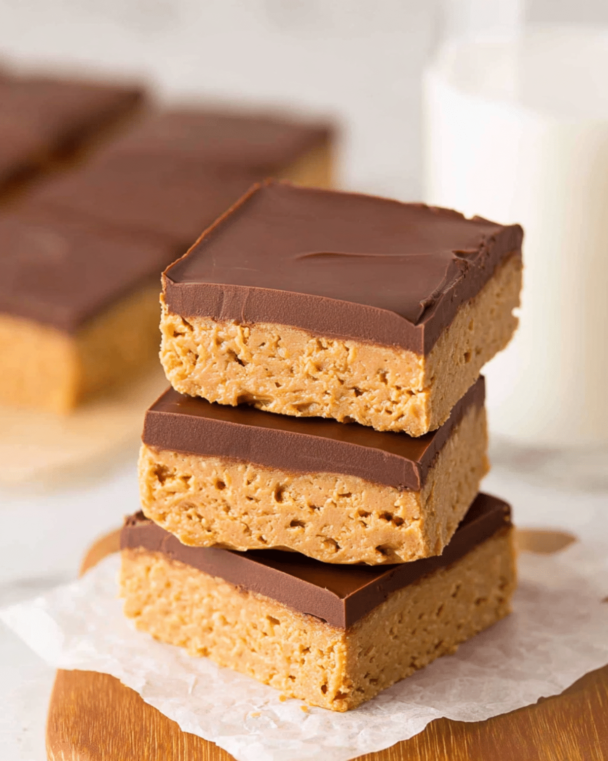 Stack of three no-bake peanut butter bars with a smooth chocolate topping on parchment paper.