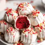 Red velvet cake balls coated in white chocolate, drizzled with icing, and topped with colorful sprinkles, displayed on a white plate.