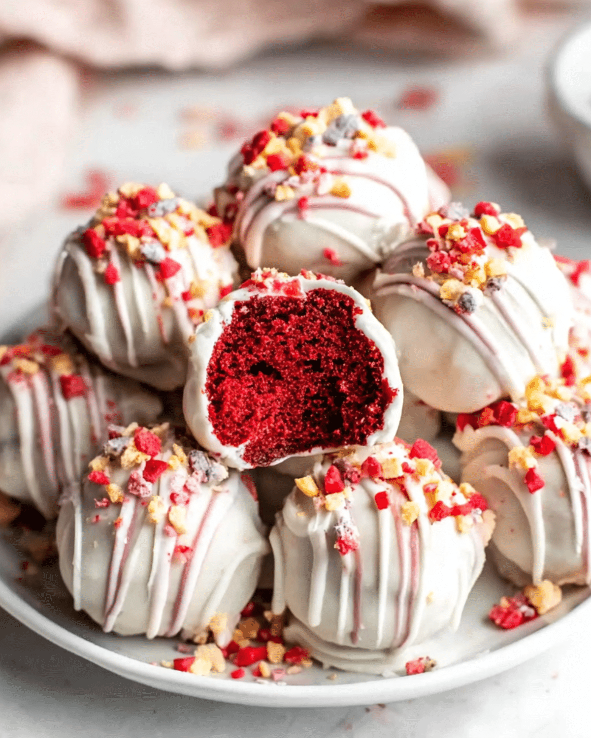 Red velvet cake balls coated in white chocolate, drizzled with icing, and topped with colorful sprinkles, displayed on a white plate.