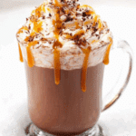 Glass mug of Salted Caramel Hot Cocoa topped with whipped cream, caramel drizzle, and chocolate shavings, sitting in snow.