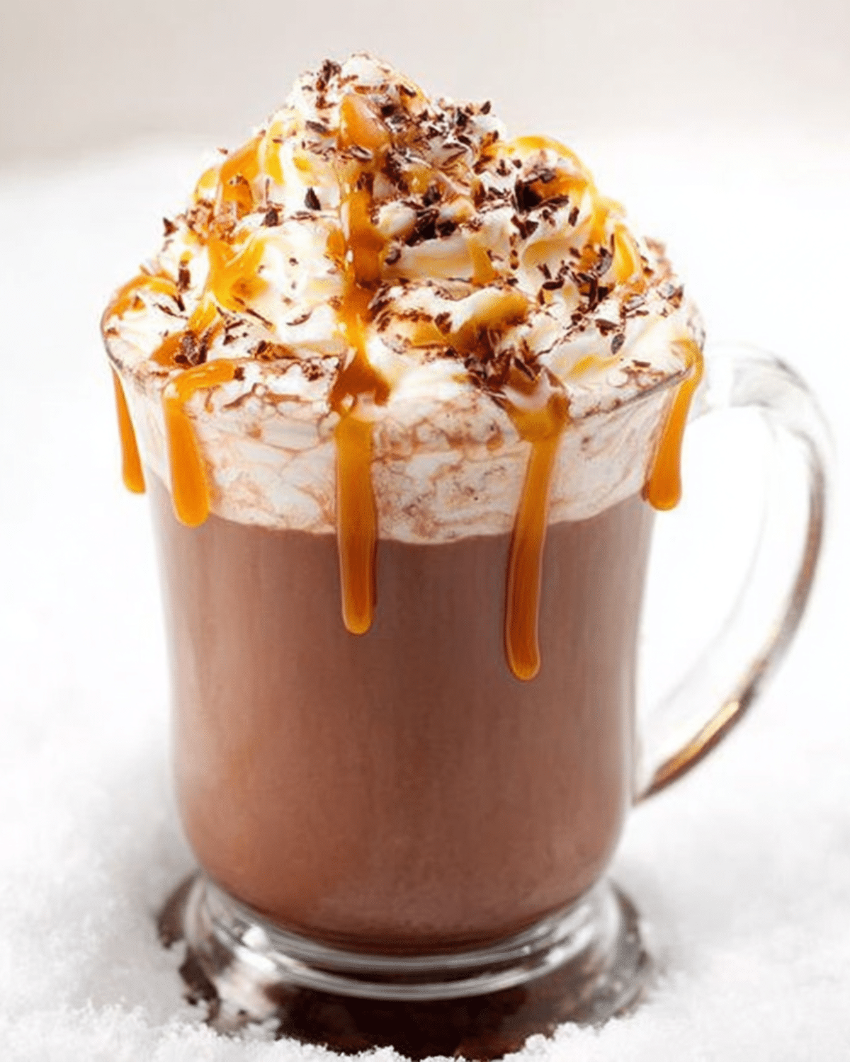 Glass mug of Salted Caramel Hot Cocoa topped with whipped cream, caramel drizzle, and chocolate shavings, sitting in snow.