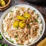 Bowl of Slow Cooker Mississippi Chicken served over mashed potatoes, topped with sliced pepperoncini and fresh herbs.
