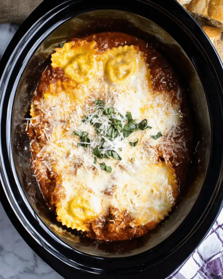 Slow Cooker Ravioli Lasagna topped with melted cheese, fresh basil, and rich marinara sauce in a black crockpot.