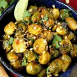 A bowl of Sweet Chili Brussel Sprouts garnished with chopped peanuts, cilantro, and a wedge of lime.