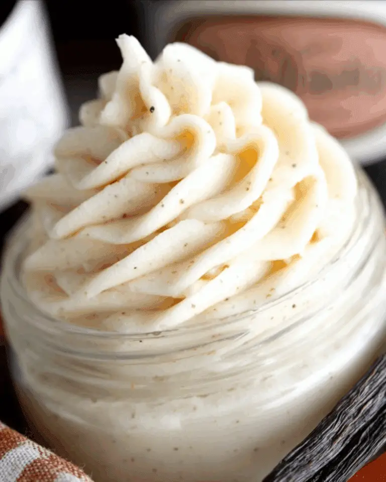 Close-up of creamy Vanilla Bean Buttercream piped into a swirl in a glass jar, with visible vanilla bean specks.