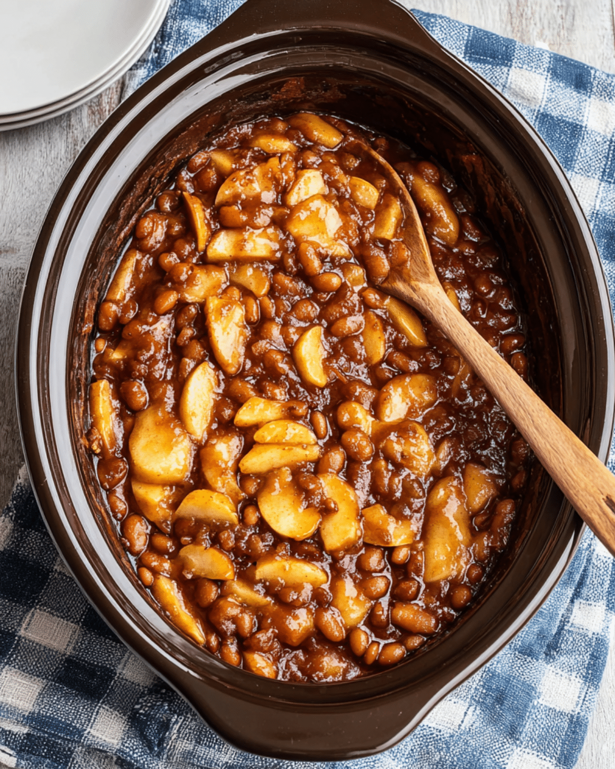 Close-up of apple pie baked beans in a slow cooker with a wooden spoon resting inside.