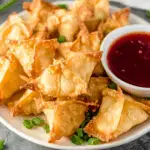 A plate of golden crispy Crab Rangoons served with a bowl of red dipping sauce and garnished with green onions.