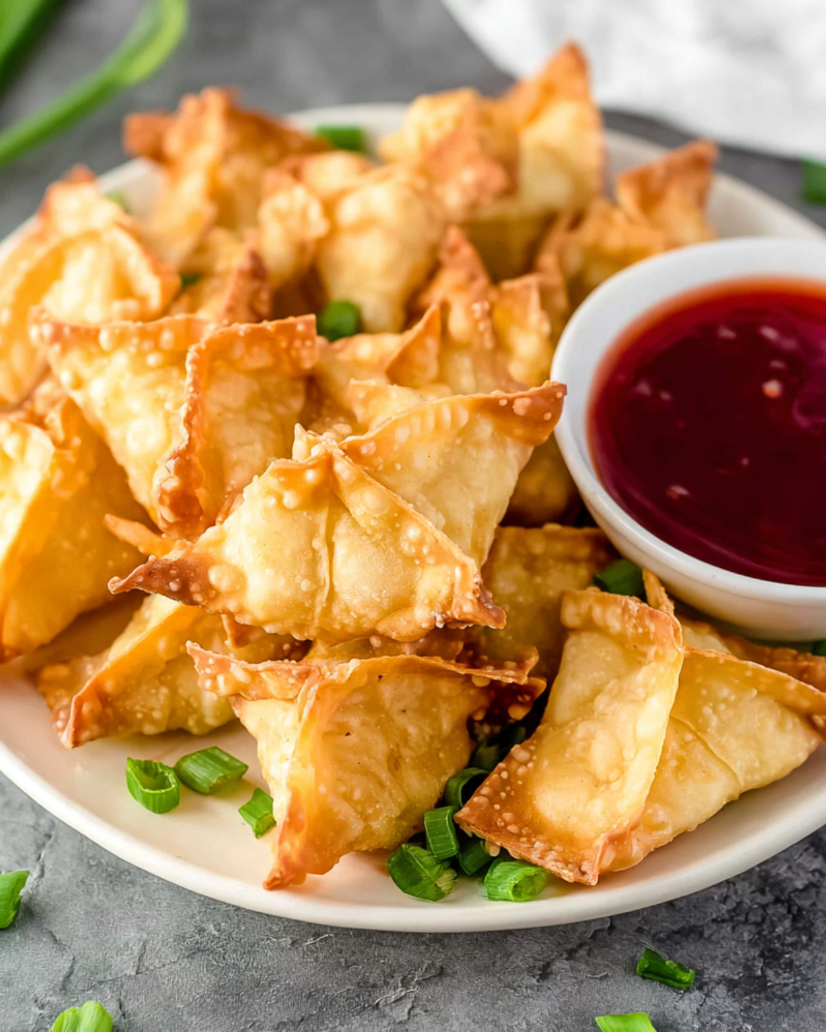A plate of golden crispy Crab Rangoons served with a bowl of red dipping sauce and garnished with green onions.