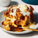 A serving of apple bread pudding topped with a rich vanilla sauce, placed on a plate with a golden fork.