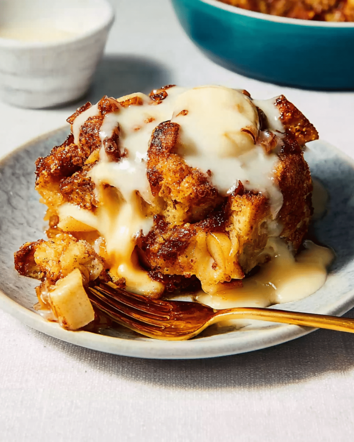 A serving of apple bread pudding topped with a rich vanilla sauce, placed on a plate with a golden fork.