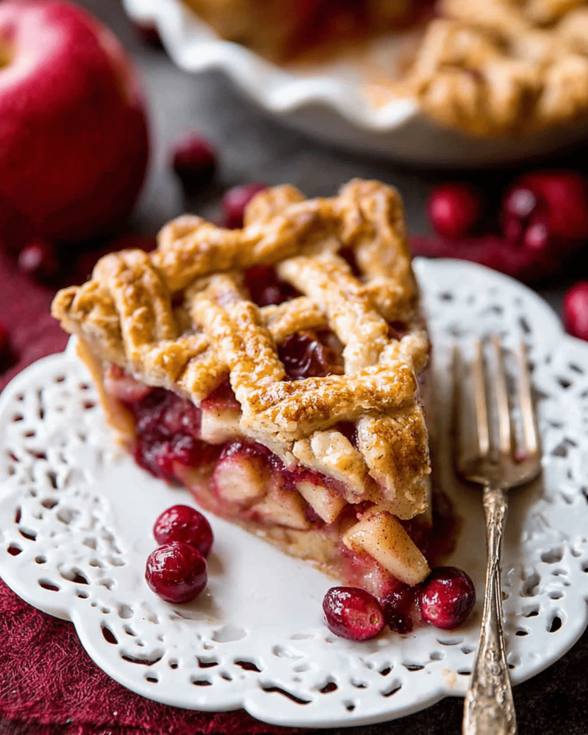 Slice of apple cranberry pie with lattice crust, served on a white plate with whole cranberries.