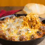 Cheesy Baked Spaghetti Meatballs in a cast iron skillet with melted cheese and marinara, served with bread.