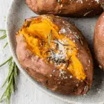 Baked sweet potato split open and topped with flaky sea salt and fresh rosemary on a speckled ceramic plate.