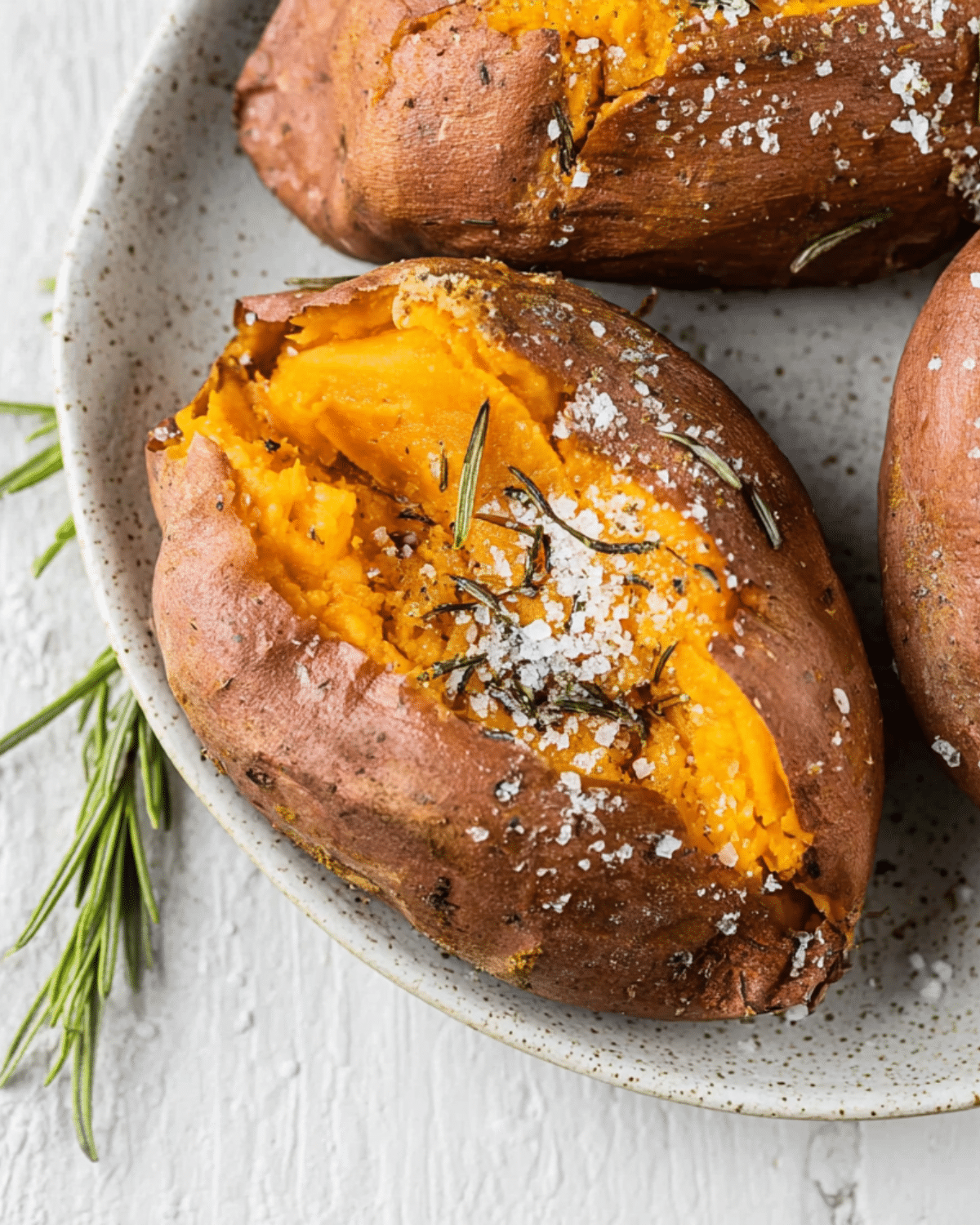 Baked sweet potato split open and topped with flaky sea salt and fresh rosemary on a speckled ceramic plate.
