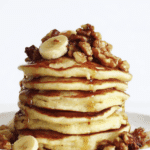 Stack of Banana Nut Pancakes topped with banana slices, glazed walnuts, and drizzled with syrup on a white plate.