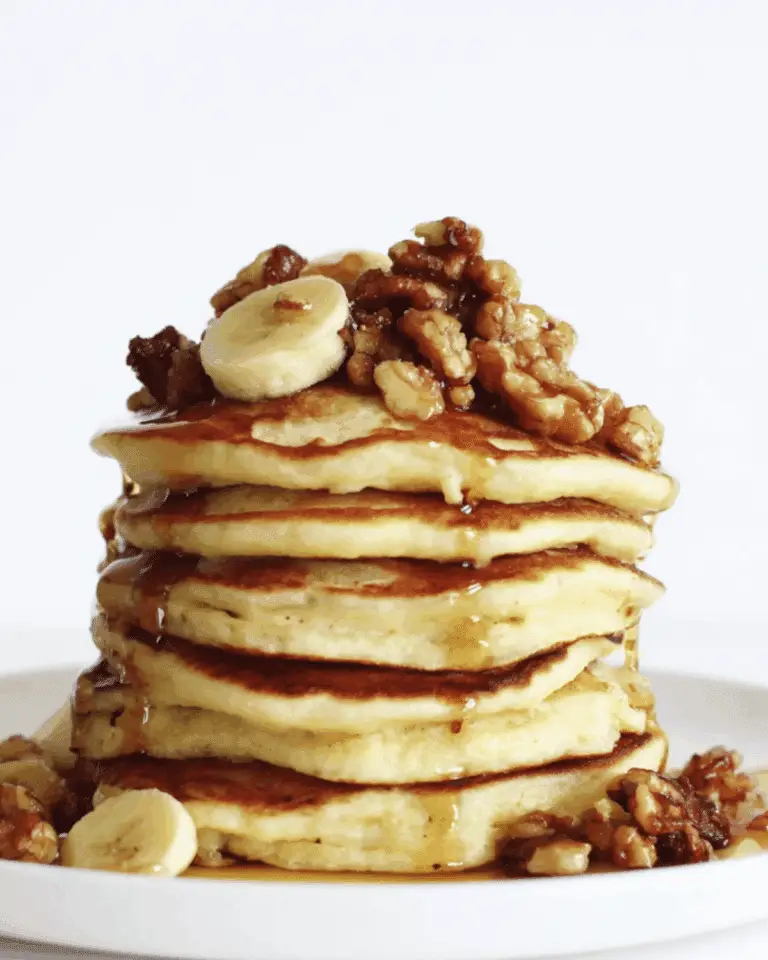Stack of Banana Nut Pancakes topped with banana slices, glazed walnuts, and drizzled with syrup on a white plate.