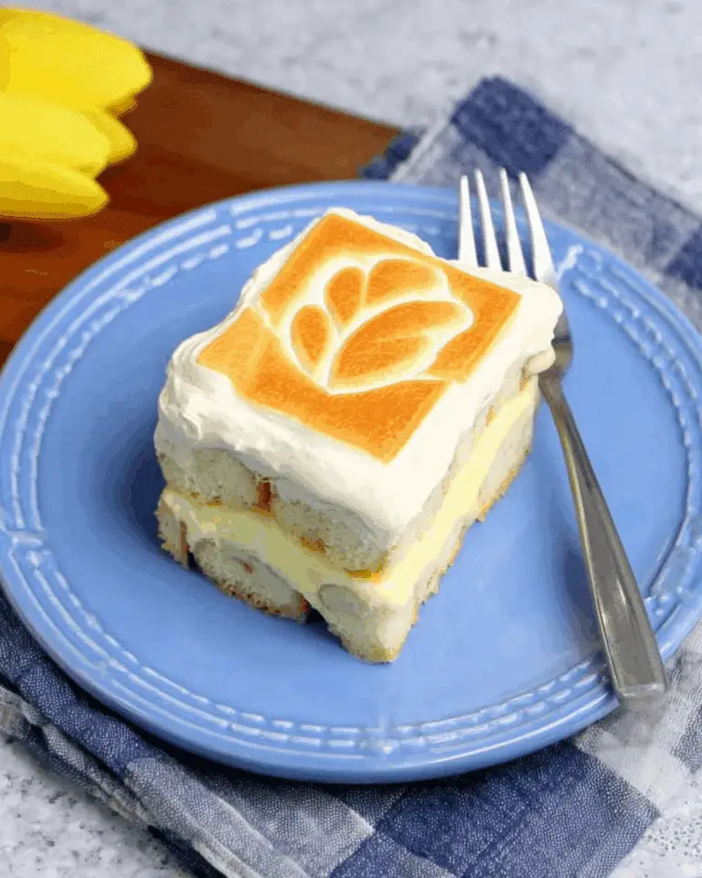 Slice of Chessman banana pudding on a blue plate, showing layers of creamy pudding, bananas, and soft cookies topped with whipped cream and a toasted design.