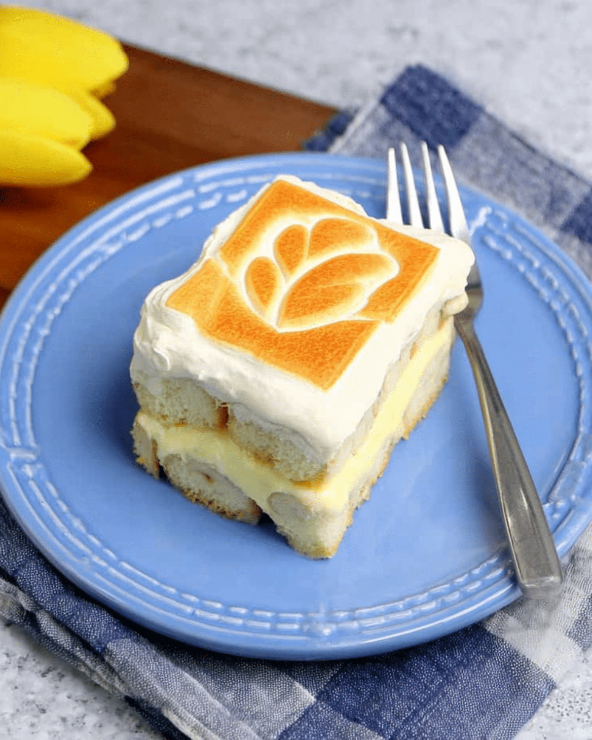 Slice of Chessman banana pudding on a blue plate, showing layers of creamy pudding, bananas, and soft cookies topped with whipped cream and a toasted design.