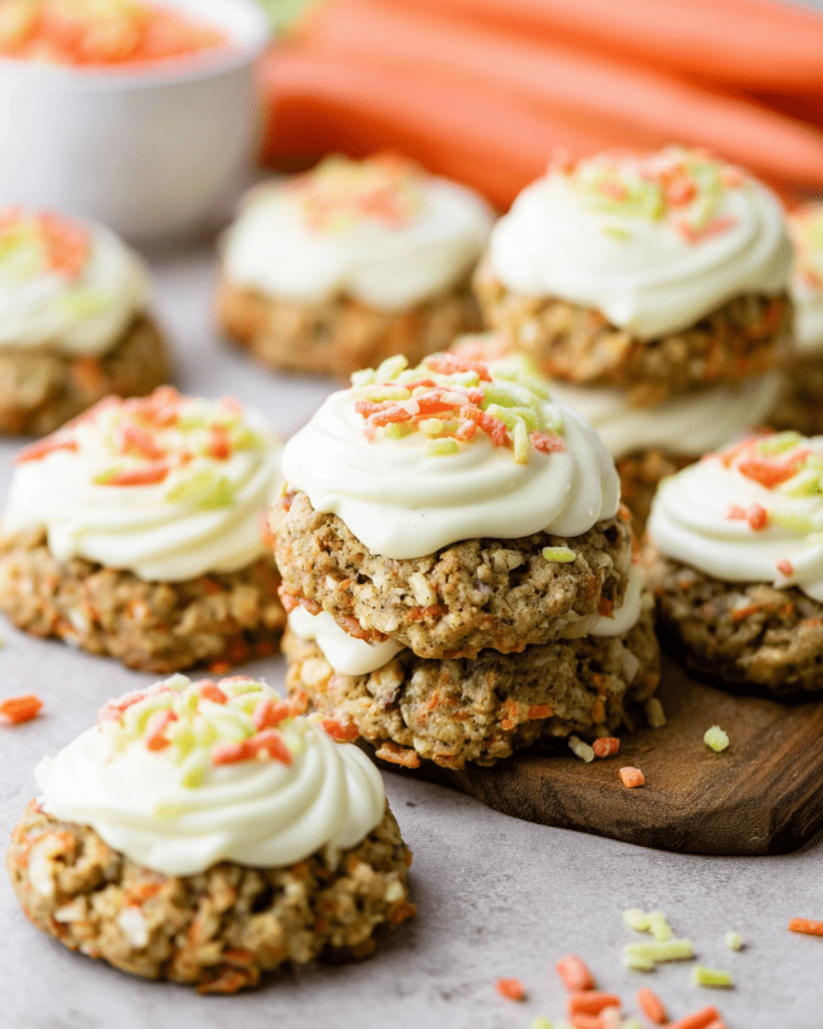 Soft carrot cake cookies topped with cream cheese frosting and colorful carrot sprinkles on a wooden board.