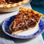 Slice of chocolate bourbon pecan pie on a blue-rimmed plate, with a rich, gooey filling and toasted pecans.