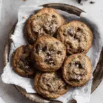 A rustic bowl lined with parchment paper filled with freshly baked chocolate chip banana cookies, topped with flaky sea salt.
