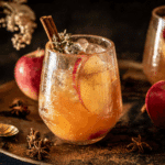 A glass of cinnamon apple bourbon smash garnished with apple slices, cinnamon sticks, and fresh thyme, surrounded by star anise and red apples.