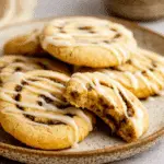 Cinnamon Roll Cookies topped with white icing drizzle, stacked on a rustic plate with one cookie bitten to reveal the soft cinnamon swirl interior.