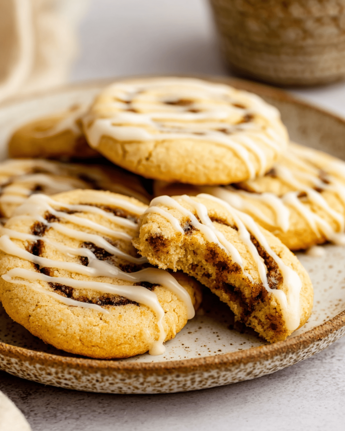 Cinnamon Roll Cookies topped with white icing drizzle, stacked on a rustic plate with one cookie bitten to reveal the soft cinnamon swirl interior.