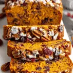 Sliced Cranberry Pecan Pumpkin Bread topped with powdered sugar, cranberries, and pecans on a white serving board.
