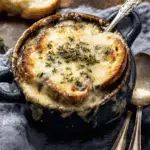 Creamy French onion and mushroom soup topped with toasted bread and melted cheese, served in a dark ceramic bowl with a spoon.