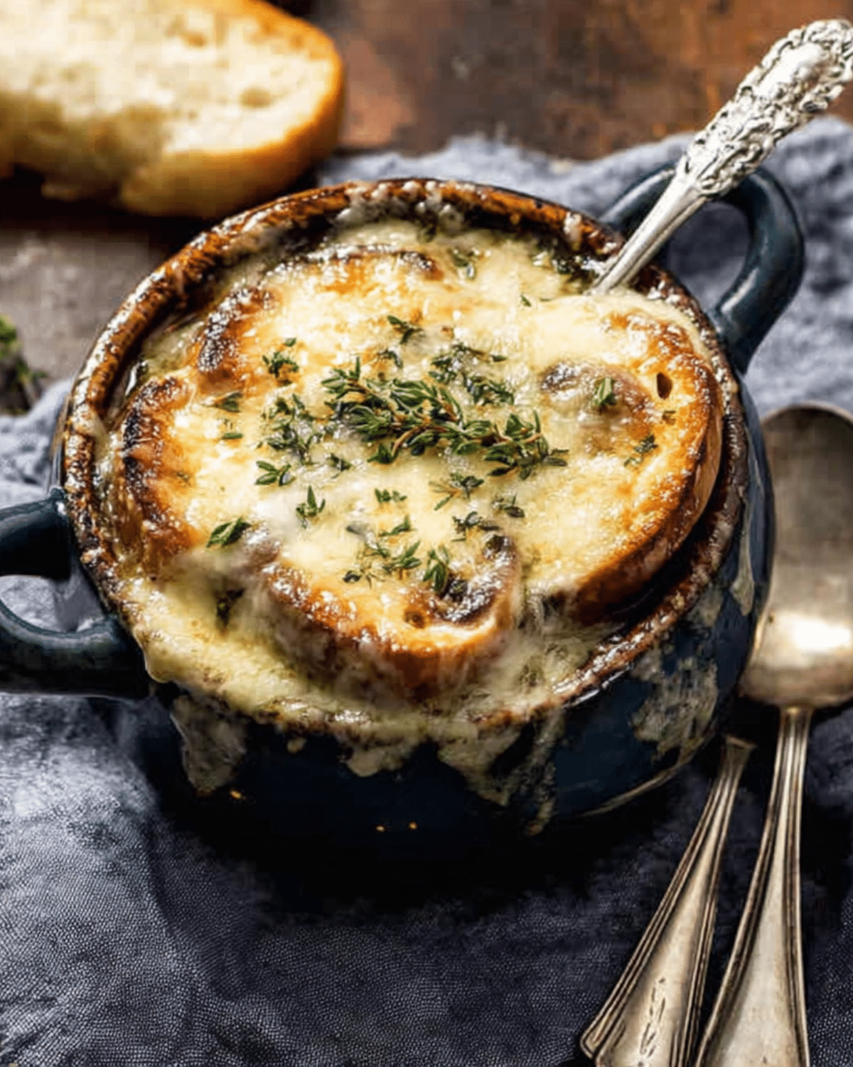 Creamy French onion and mushroom soup topped with toasted bread and melted cheese, served in a dark ceramic bowl with a spoon.