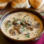 Creamy mushroom soup garnished with fresh parsley in a brown ceramic bowl, served with crusty bread rolls in the background.