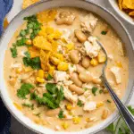 A bowl of creamy white chicken chili filled with shredded chicken, white beans, corn, and garnished with cilantro and crushed tortilla chips.
