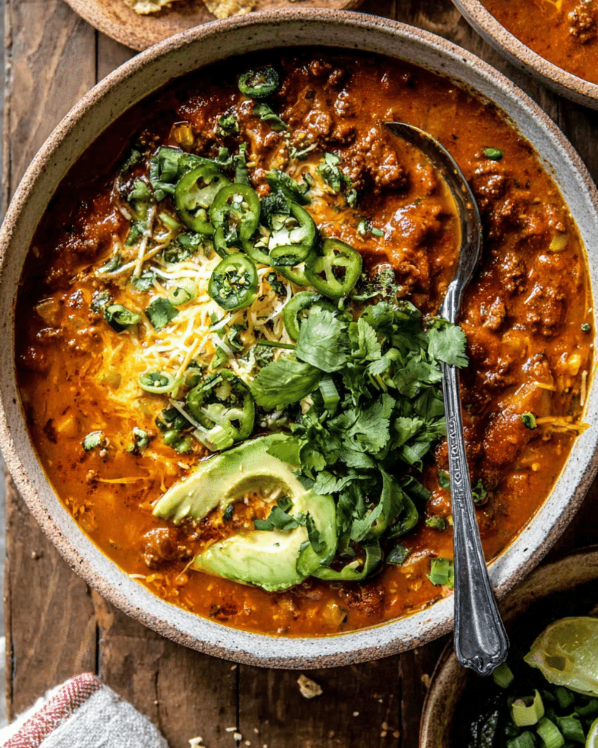 A bowl of crockpot spicy queso beef chili topped with shredded cheese, sliced jalapeños, avocado, and fresh cilantro.
