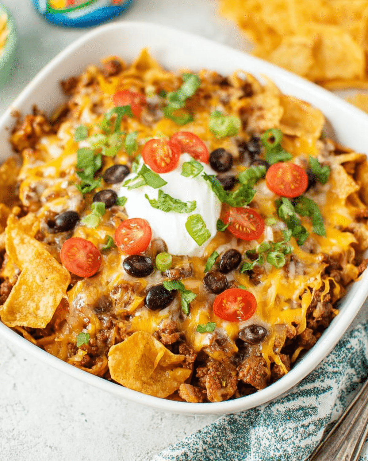 Frito Pie Casserole topped with melted cheese, black beans, cherry tomatoes, green onions, and sour cream in a white dish.