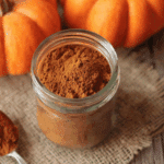 A glass jar filled with homemade pumpkin pie spice surrounded by fresh pumpkins and a spoonful of spice on a rustic burlap cloth.