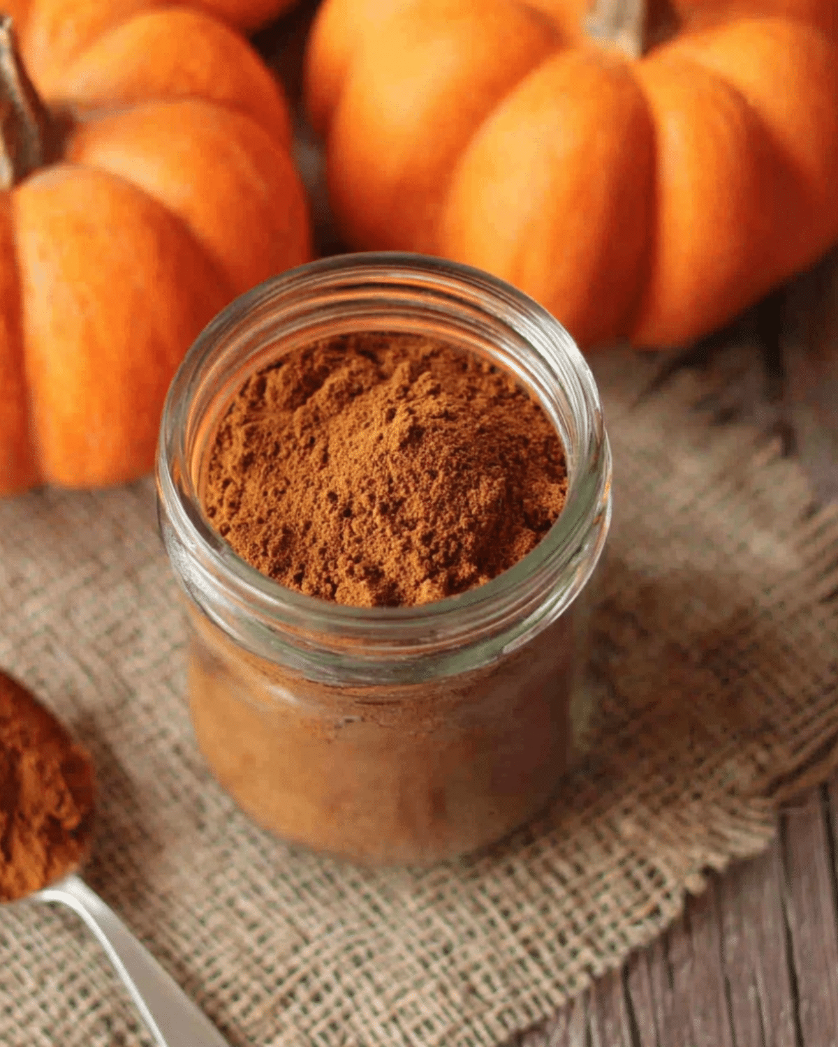 A glass jar filled with homemade pumpkin pie spice surrounded by fresh pumpkins and a spoonful of spice on a rustic burlap cloth.