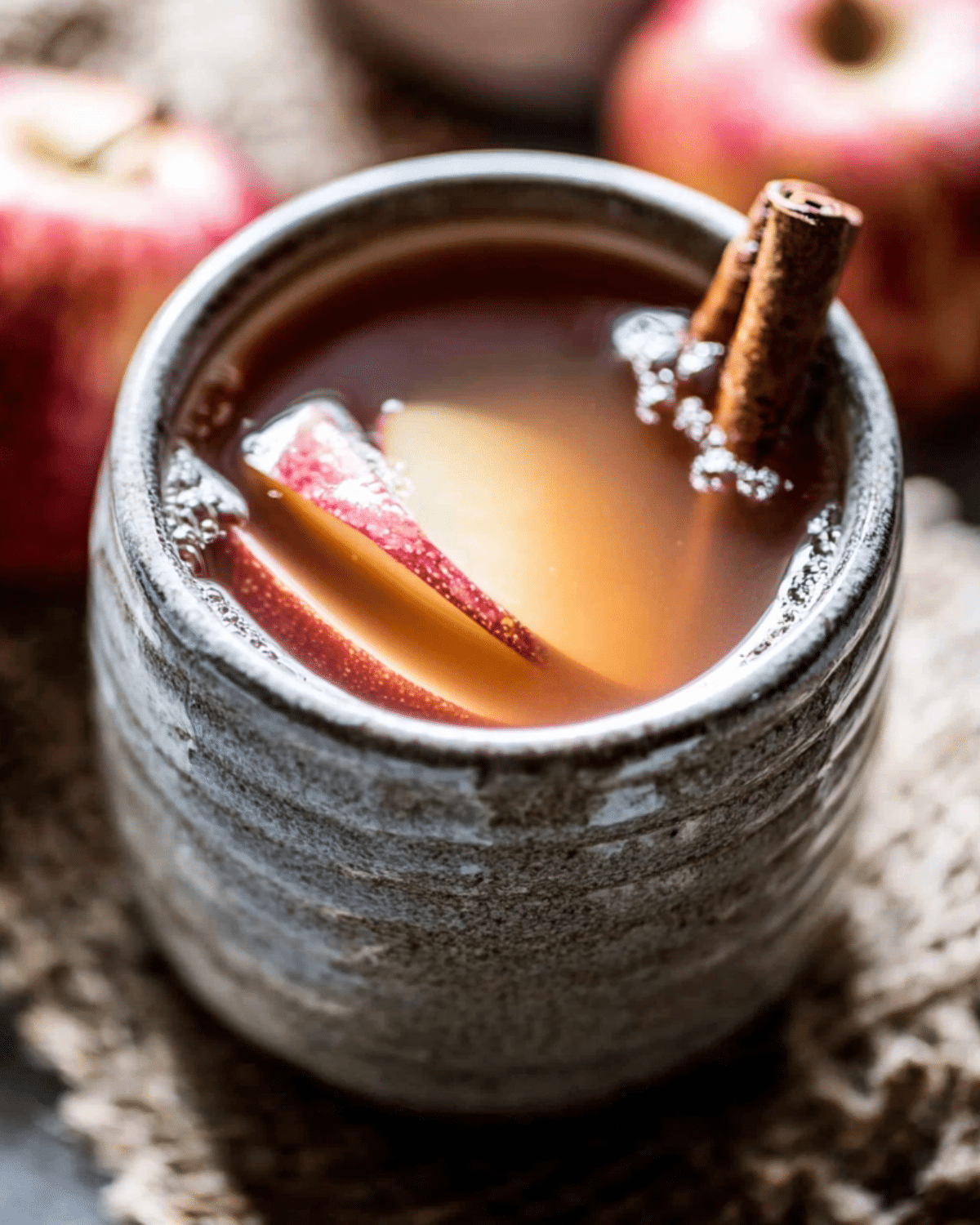 Close-up of a ceramic mug filled with maple apple cider, garnished with apple slices and cinnamon sticks.