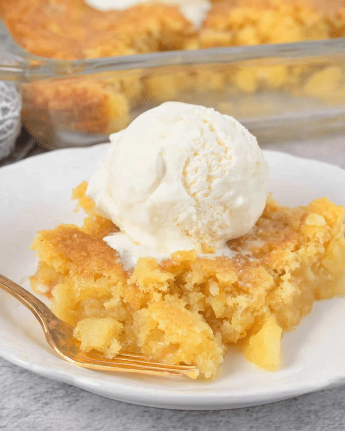 A serving of pineapple dump cake topped with a scoop of vanilla ice cream on a white plate with a golden fork.