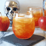 Chilled glass of Poisoned Apple Cider with ice, surrounded by red apples and Halloween-themed decor, including a silver skull.