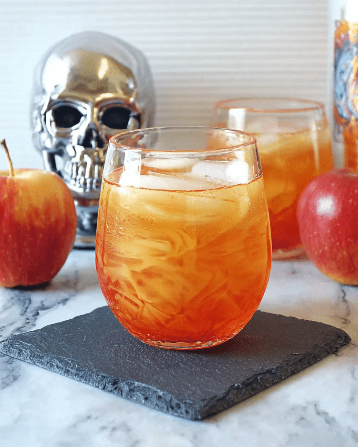 Chilled glass of Poisoned Apple Cider with ice, surrounded by red apples and Halloween-themed decor, including a silver skull.