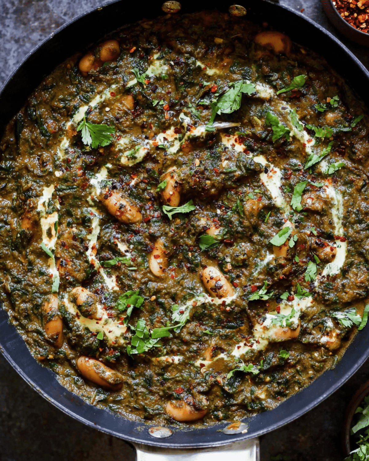 A skillet filled with saag butter beans, garnished with fresh cilantro, cream swirls, and red chili flakes.