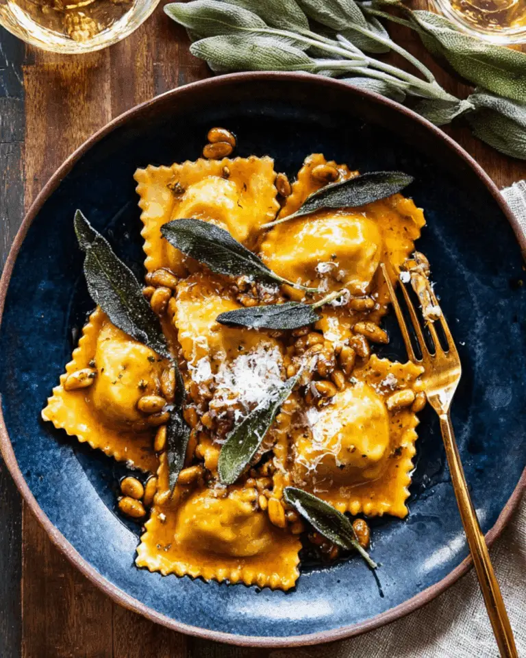 Sage butter pumpkin cheese ravioli served on a dark blue ceramic plate, topped with crispy sage leaves, toasted pine nuts, and grated cheese.