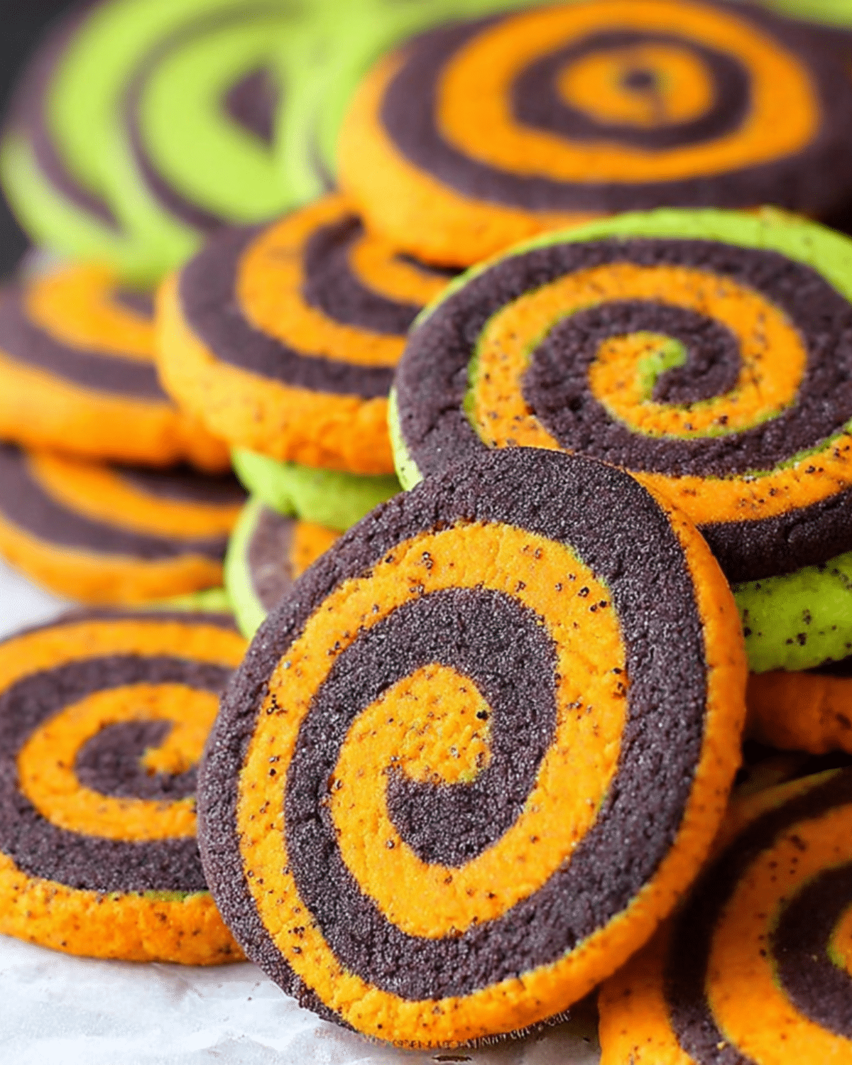 Bright orange and black spiral Halloween cookies stacked together, with a few green-accented cookies in the background.