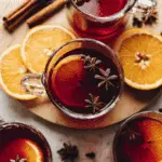Glass mugs filled with tart cherry mulled drink, garnished with orange slices and star anise, surrounded by cinnamon sticks and fresh orange slices.