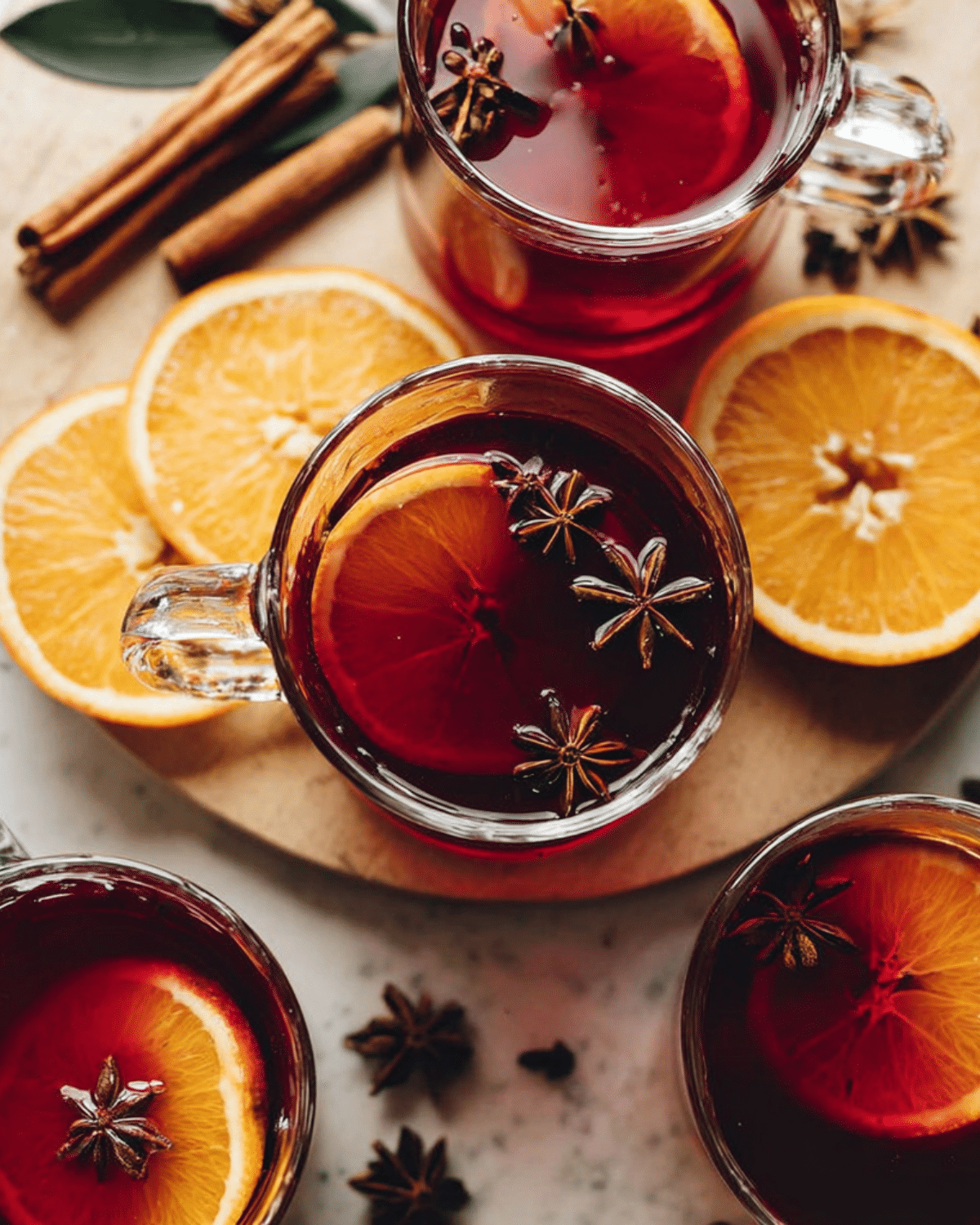 Glass mugs filled with tart cherry mulled drink, garnished with orange slices and star anise, surrounded by cinnamon sticks and fresh orange slices.