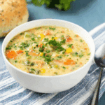 Veggie pot pie soup in a white bowl, filled with carrots, corn, peas, and herbs, served with a seeded bread roll on the side.