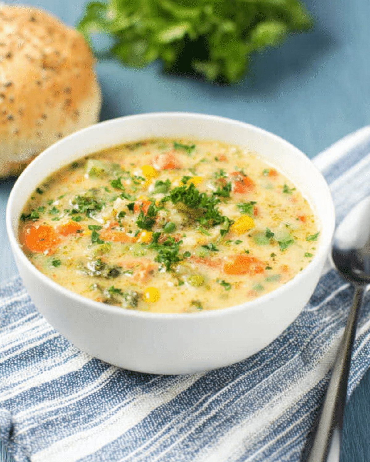 Veggie pot pie soup in a white bowl, filled with carrots, corn, peas, and herbs, served with a seeded bread roll on the side.