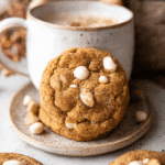 White chocolate chai pumpkin snickerdoodle cookie leaning against a ceramic mug filled with a frothy drink.